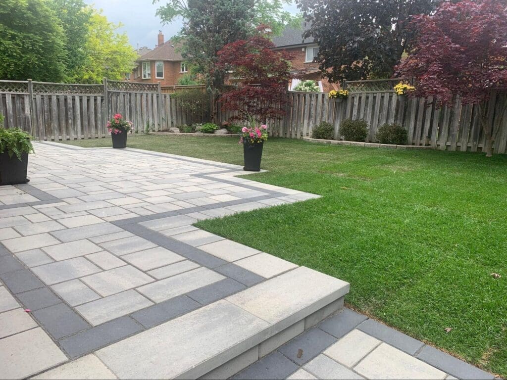 Spacious backyard patio with light interlocking stones, outdoor dining area and modern wicker furniture surrounded by a green lawn.